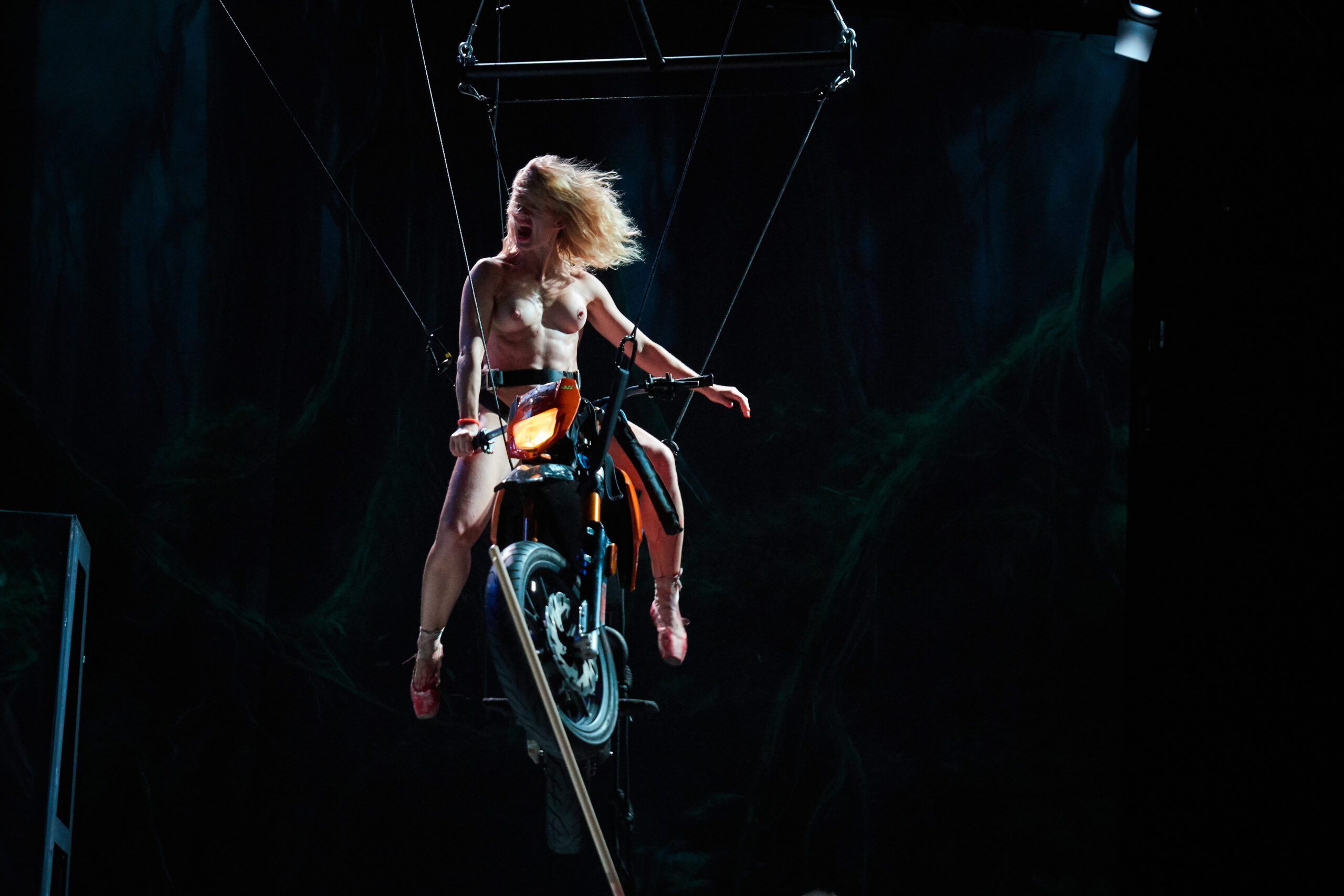 A person in minimal clothing, wearing ballet shoes, is suspended on a motorcycle by wires on a dark stage, with their hair flowing as they look down the ramp below.