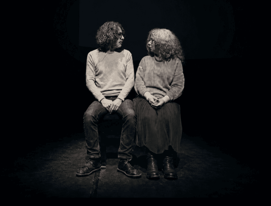 Two people with curly hair and glasses sit side by side on a dark stage, facing each other and making eye contact. Both wear sweaters and look serious or focused.