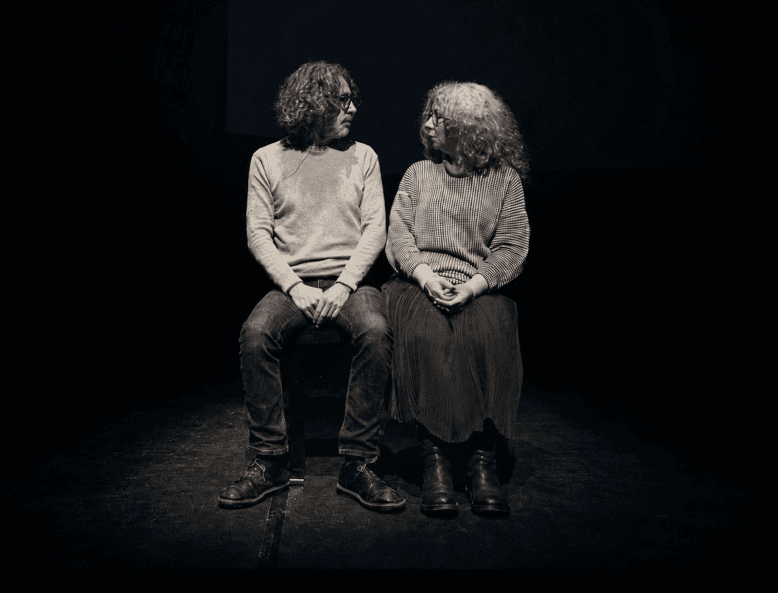 Two people with curly hair and glasses sit side by side on a dark stage, facing each other and making eye contact. Both wear sweaters and look serious or focused.