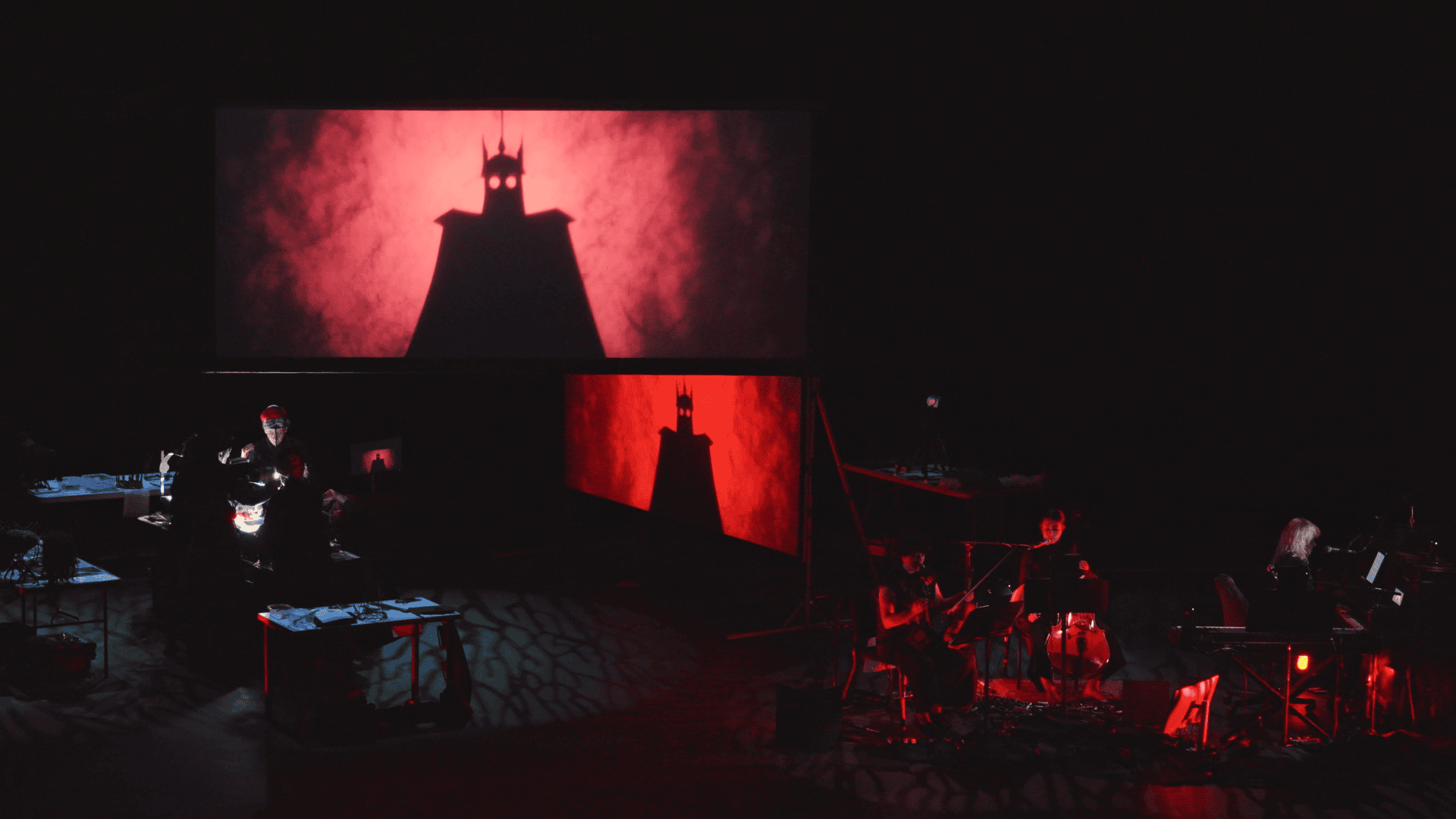 A stage with musicians performing beside a screen showing a shadowy figure with pointed ears and a red, smoky background, evoking a dramatic atmosphere.