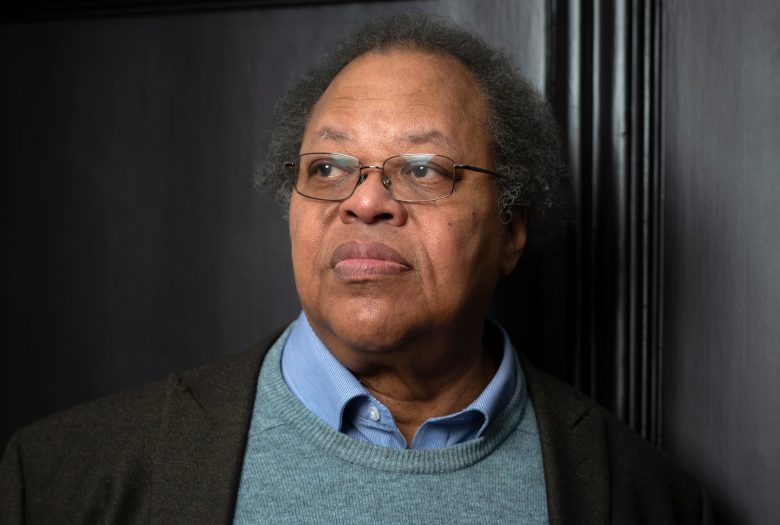 An older man with glasses, gray hair, and a thoughtful expression stands in front of a dark wall, wearing a blue shirt, green sweater, and brown blazer.
