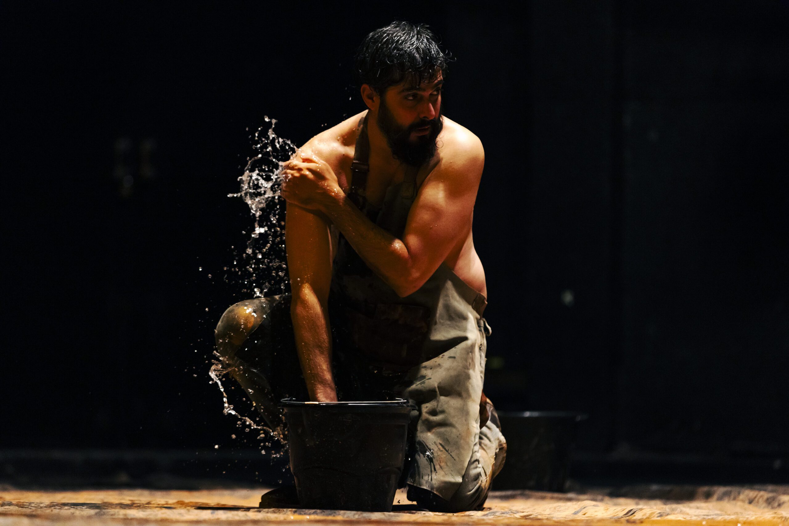 A bearded man kneels shirtless on the ground, splashing water from a black bucket onto his shoulder, with a dark background and dramatic lighting highlighting his muscles.