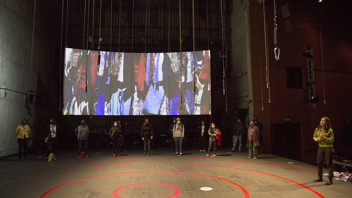 A group of people stand spaced out in a dark theater, facing a large curved screen showing abstract visuals. Red circles are marked on the floor, and ropes or cables hang from above.