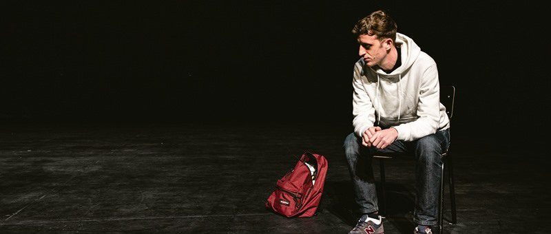A man in a white hoodie sits on a chair on a dark stage, looking down thoughtfully, with a red backpack on the floor beside him.