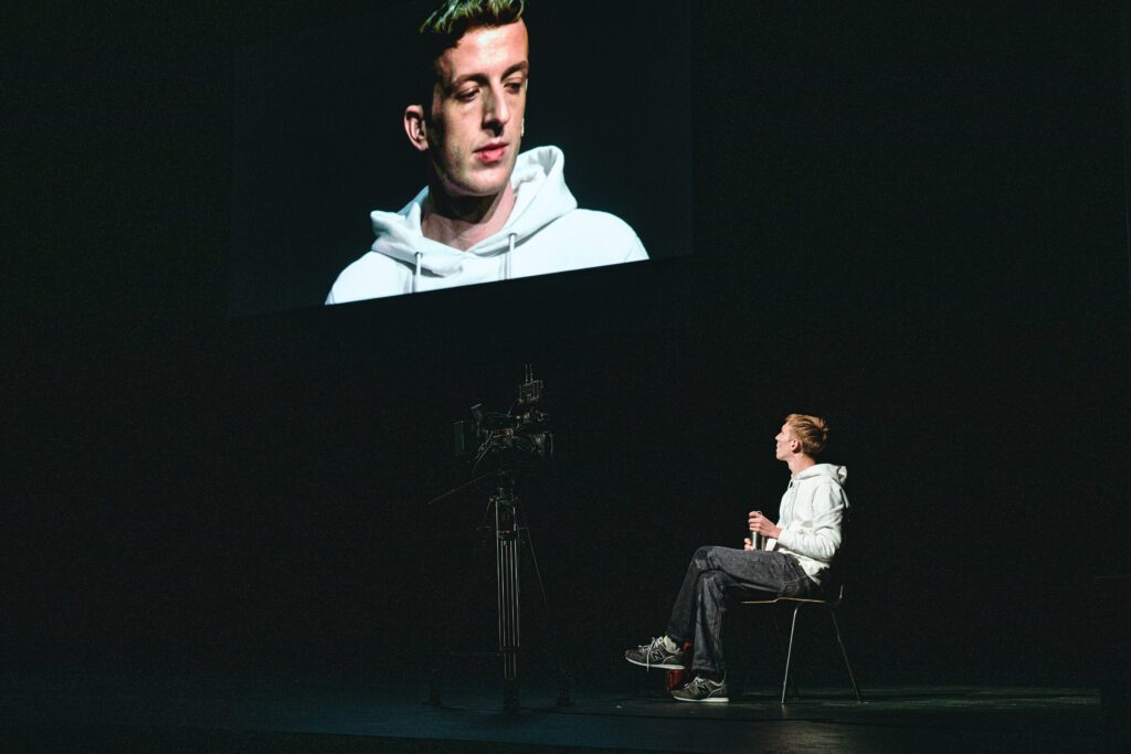 A person in a white hoodie sits on a chair facing a camera, with their large image projected on a screen above in a dark room.