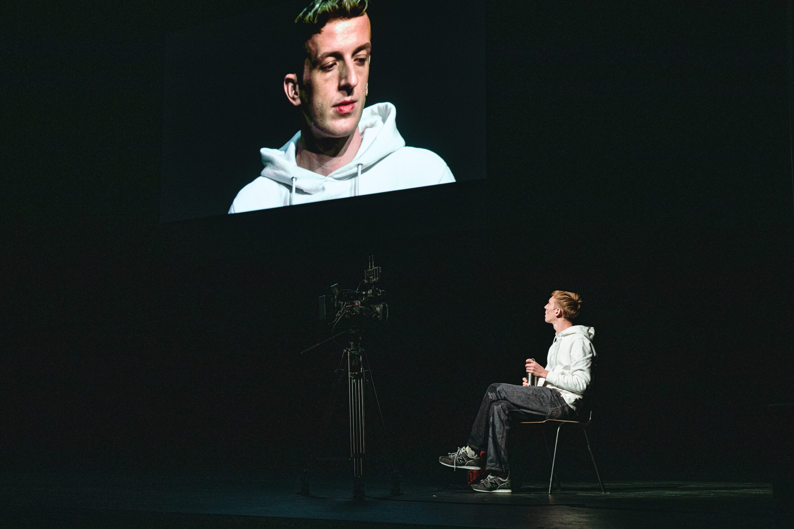A person in a white hoodie sits on a chair facing a camera, with their large image projected on a screen above in a dark room.