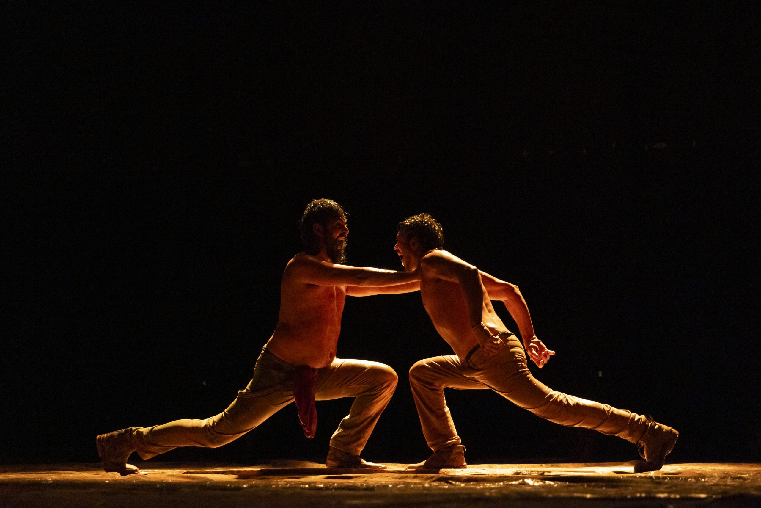 Two men in beige pants, shirtless, face each other in a dramatic, low-lit dance pose with outstretched arms, appearing to balance and resist each other on a dimly lit stage.