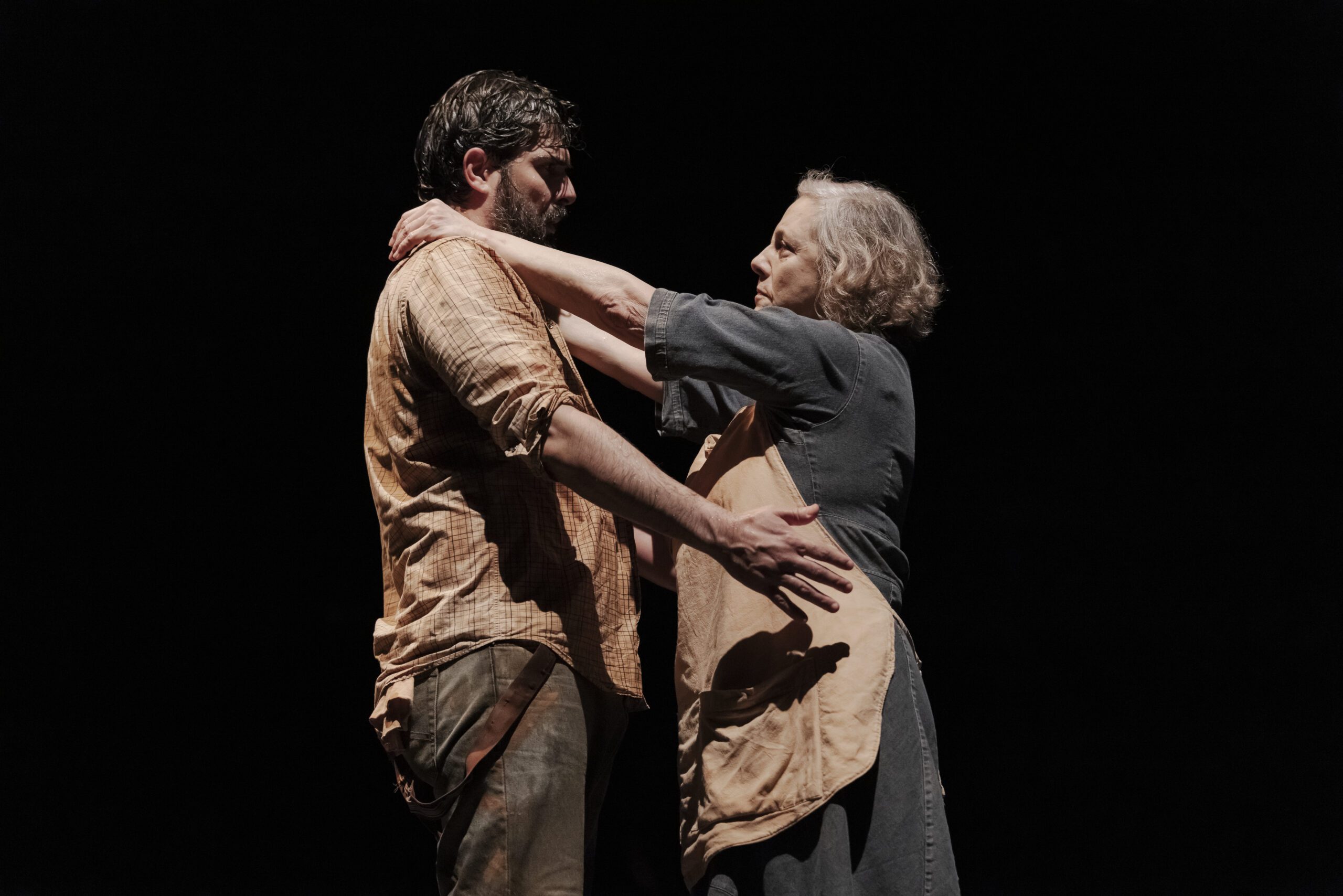 An older woman gently holds a man’s shoulders and waist. They face each other with serious expressions against a dark, plain background.