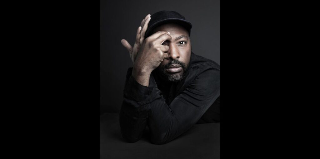 A person in dark clothing and a cap poses against a dark background, resting an arm on a surface and placing a hand on their forehead, gazing directly at the camera with a thoughtful expression.