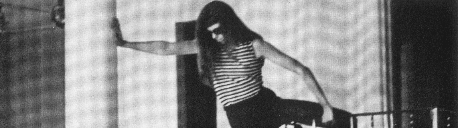 A person in a striped shirt balances on a railing, holding a column for support, in a black-and-white indoor scene.