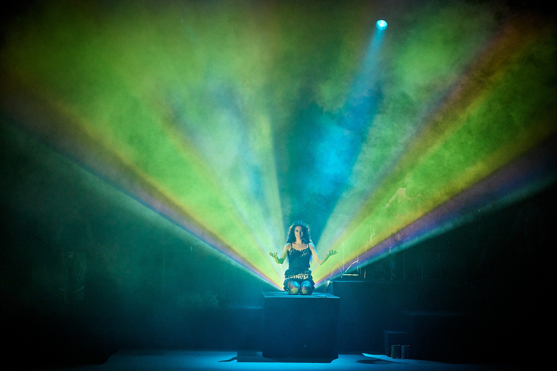 A performer kneels on a platform with arms open, surrounded by colorful beams of light and mist, creating a dramatic, vibrant stage effect.
