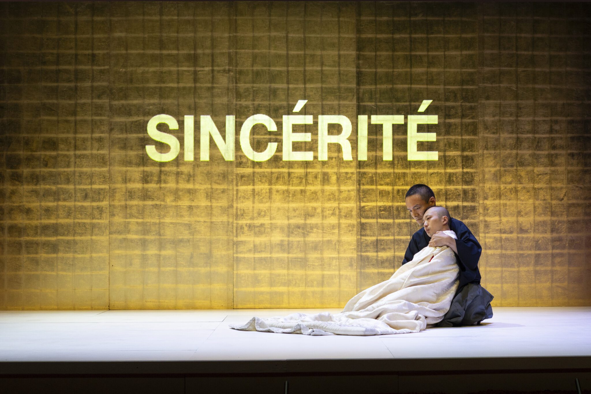 Two people sit on a stage under a blanket, one comforting the other. The word “SINCÉRITÉ” is illuminated in large letters on a golden, textured background behind them.