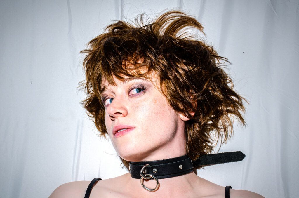 Person with tousled brown hair wearing a black collar with an O-ring, looking confidently at the camera against a white background.