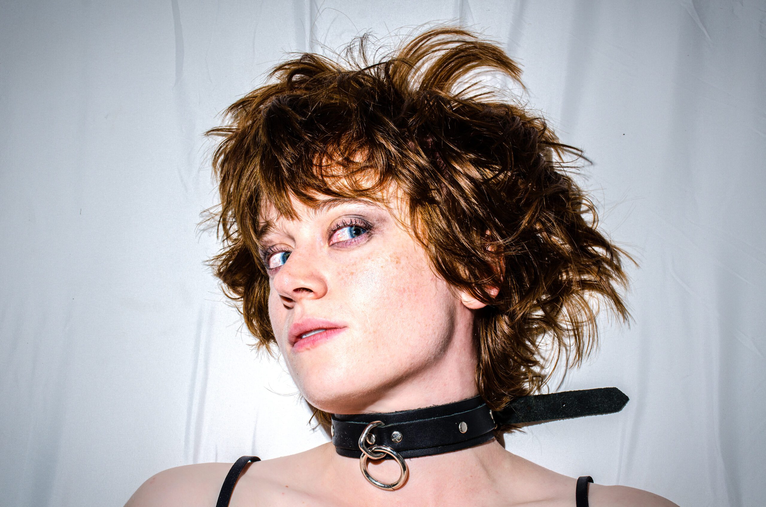 Person with tousled brown hair wearing a black collar with an O-ring, looking confidently at the camera against a white background.