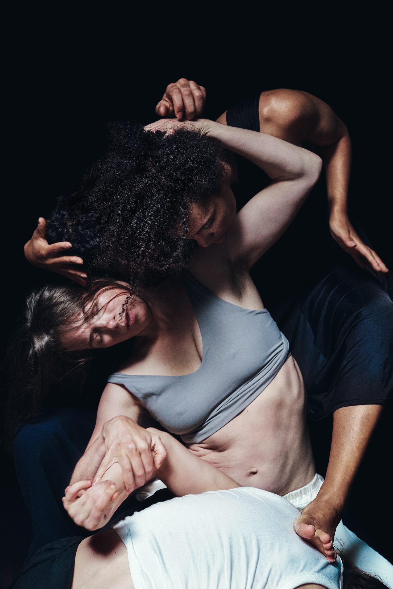 Three dancers intertwine their bodies in a complex, expressive pose against a dark background, showcasing athleticism and connection through their overlapping arms, legs, and focused expressions.