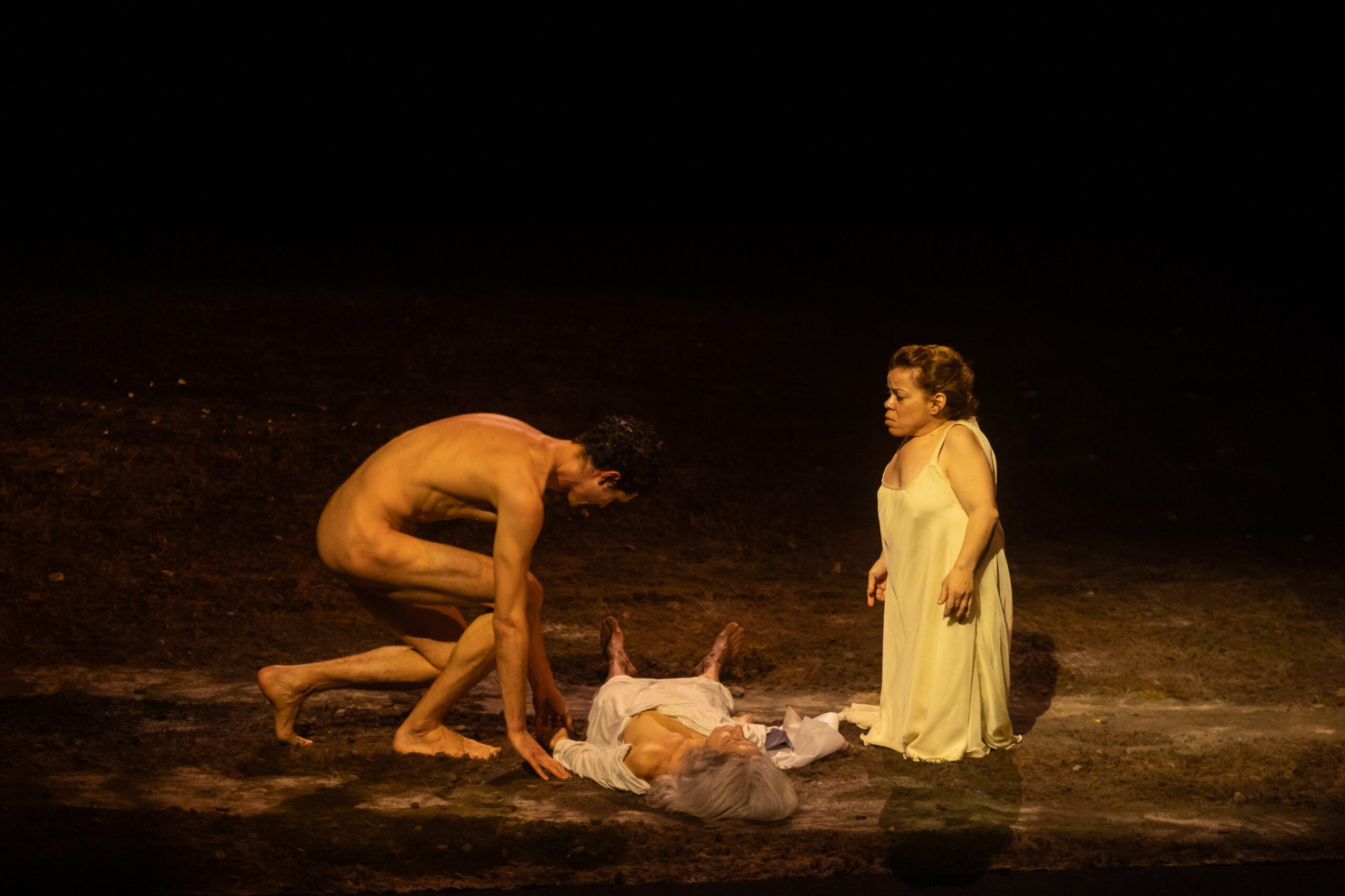 A naked man bends over a person lying on the ground covered in a white cloth, while a woman in a pale dress stands nearby, all under dramatic stage lighting.