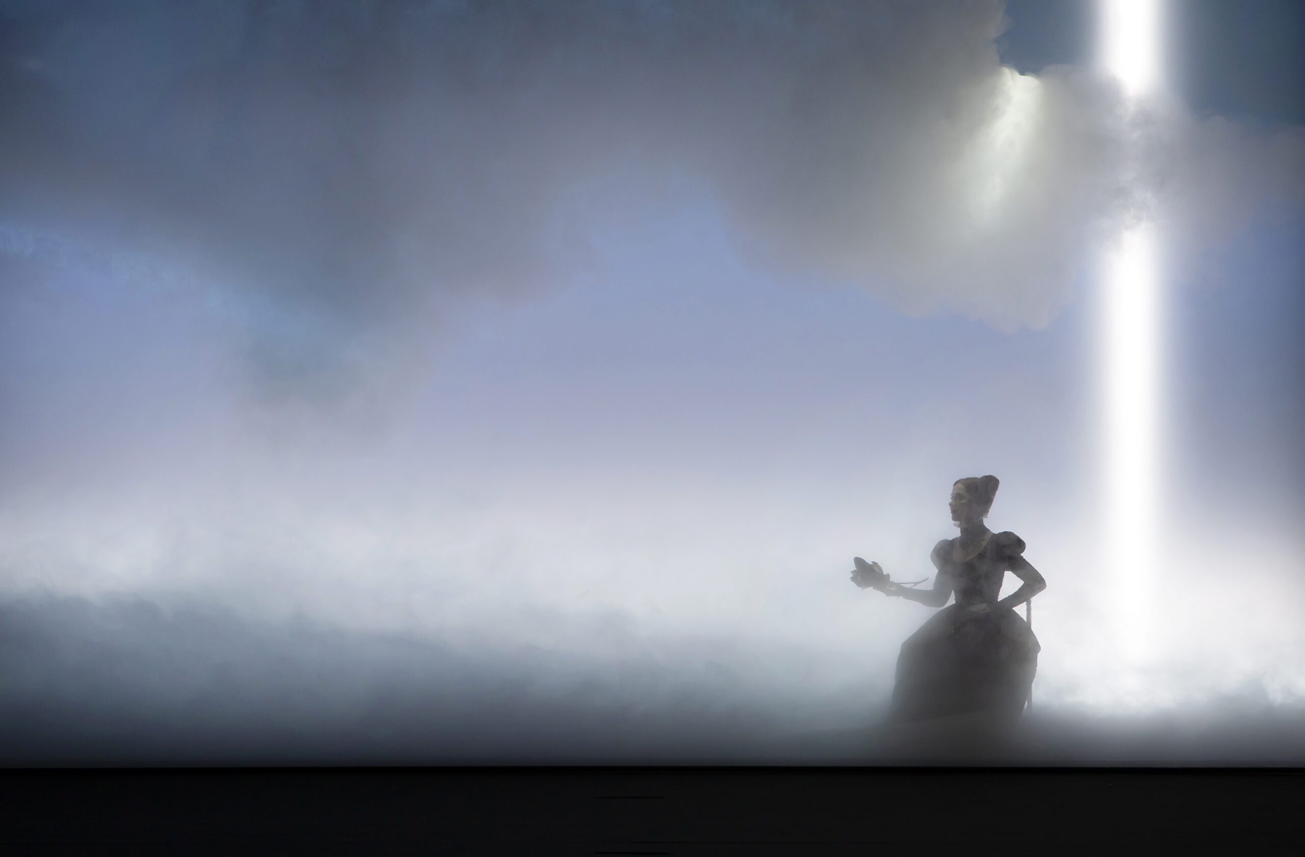 A woman in a long dress stands in fog with dramatic clouds above and a bright beam of light shining down near her.