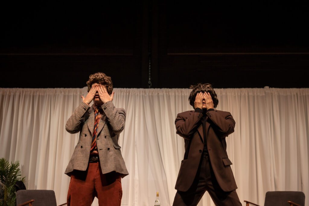 Two people in suits stand side by side onstage, both covering their eyes with their hands. There is a white curtain backdrop, chairs, and a small plant on the left side.