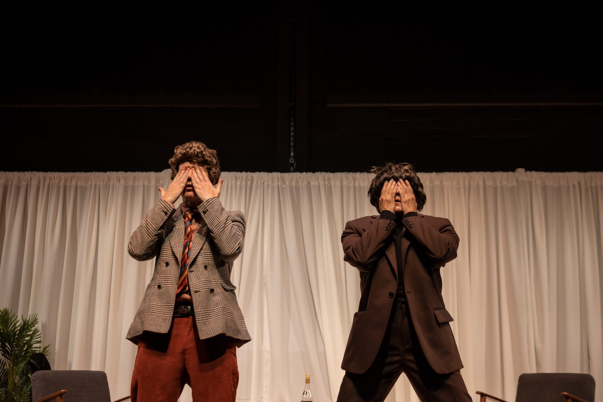 Two people in suits stand side by side onstage, both covering their eyes with their hands. There is a white curtain backdrop, chairs, and a small plant on the left side.