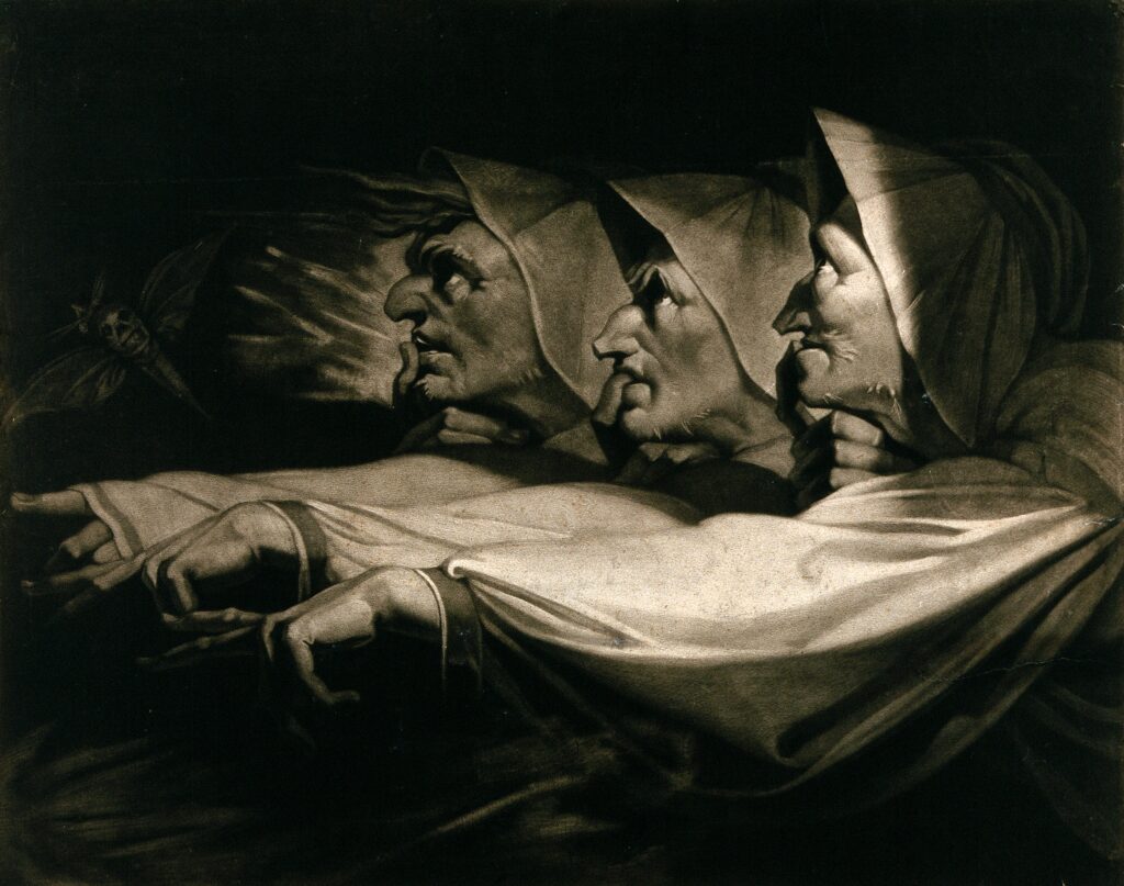 Three women in hooded cloaks with exaggerated features point forward dramatically, their faces lit from below, creating deep shadows and a mysterious, ominous atmosphere.