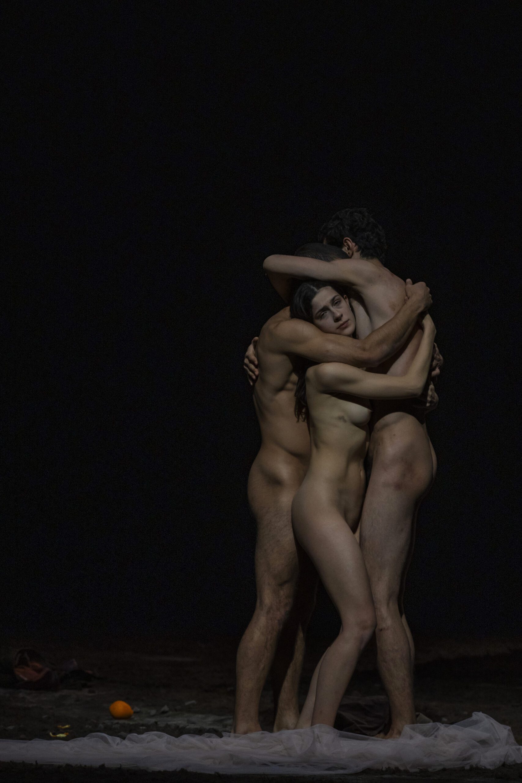 Three nude individuals stand closely together in a gentle embrace on a dimly lit stage, with one person resting their head on another’s shoulder. A white sheet and an orange object are on the floor nearby.