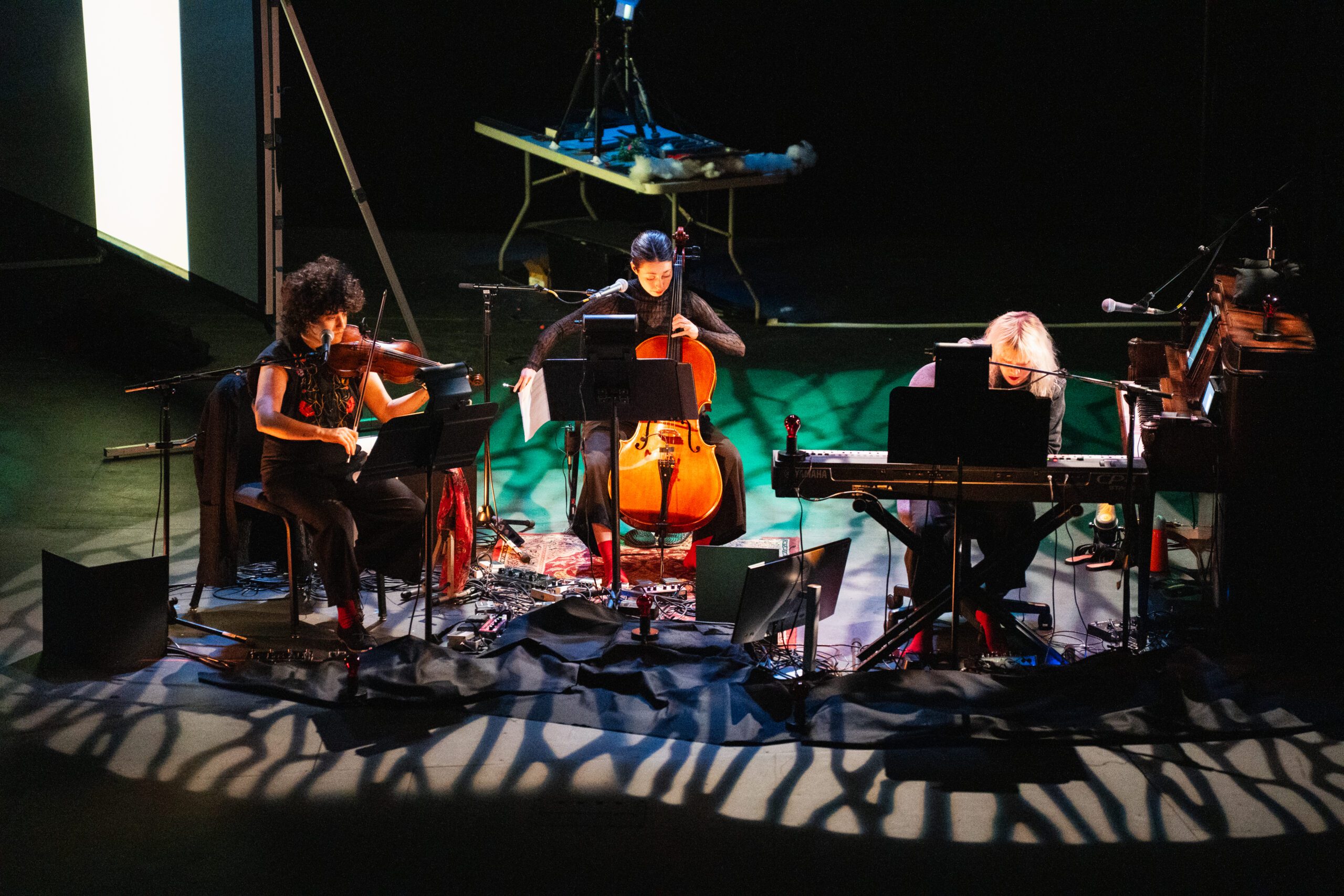 Three musicians perform on stage with a cello, violin, and keyboard. Music stands and equipment surround them, while patterned lighting casts shadows across the floor. The background is dark and a table holds technical gear.
