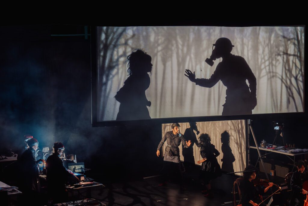 A theater performance with actors in front of a screen showing their silhouettes; one silhouette wears a gas mask. Musicians perform on the left side, surrounded by equipment and dramatic lighting.
