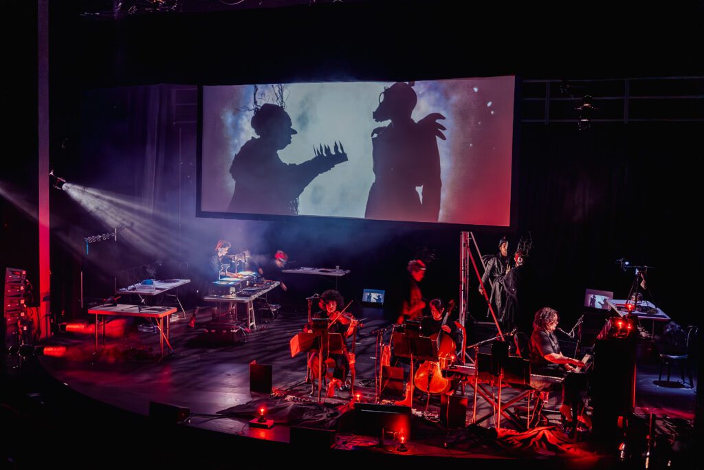 A stage performance features musicians playing string instruments below a screen displaying a dramatic shadow scene with two figures. Red and purple lighting creates a moody, theatrical atmosphere.