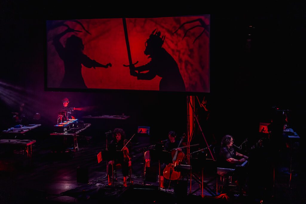 A stage performance features musicians playing with a large screen above them showing dramatic red and black shadow puppets of two figures, one holding a sword, set against an eerie backdrop.