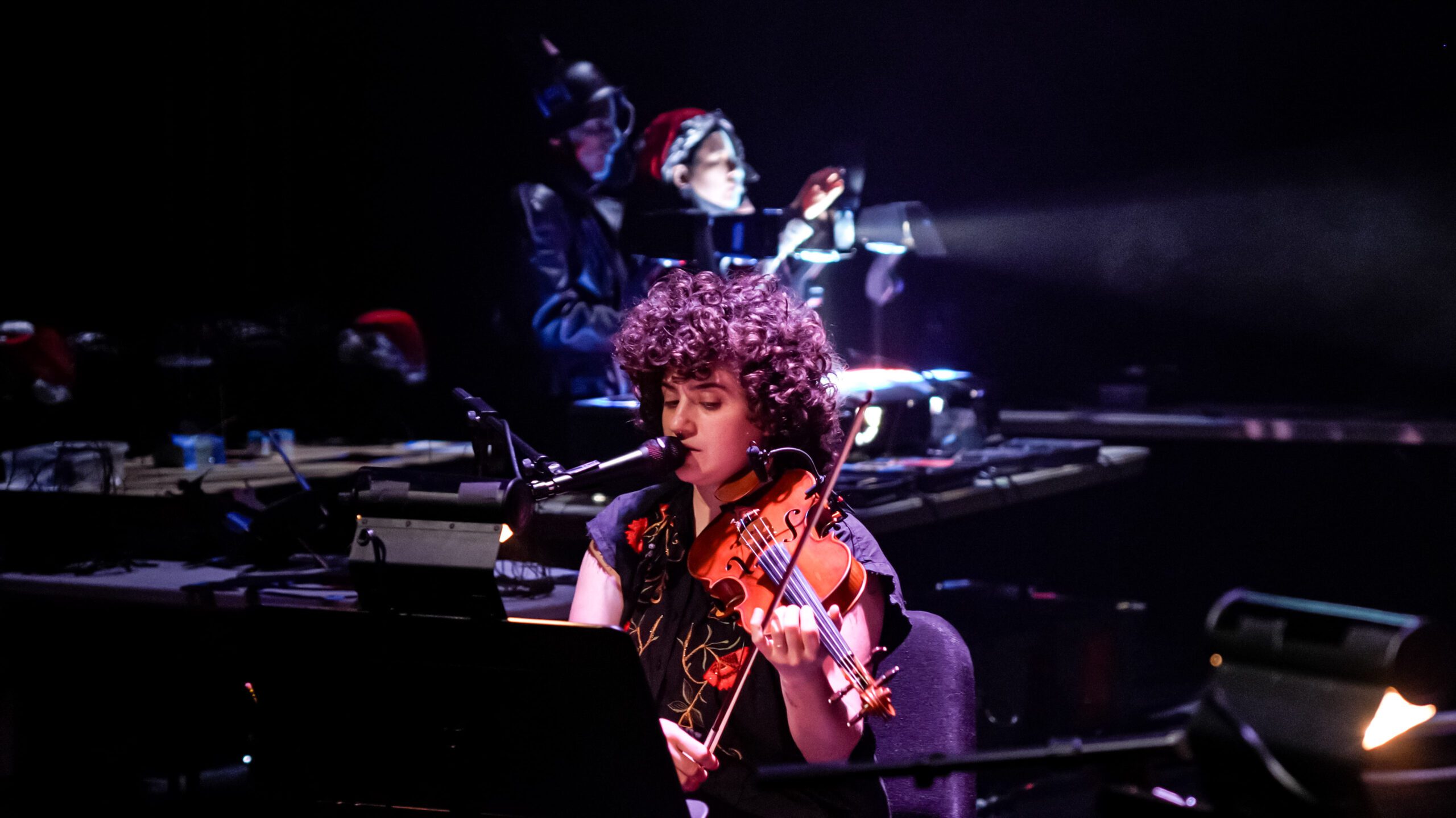 A musician with curly hair sings into a microphone while playing a violin on stage. In the background, two people operate electronic equipment under dramatic lighting.