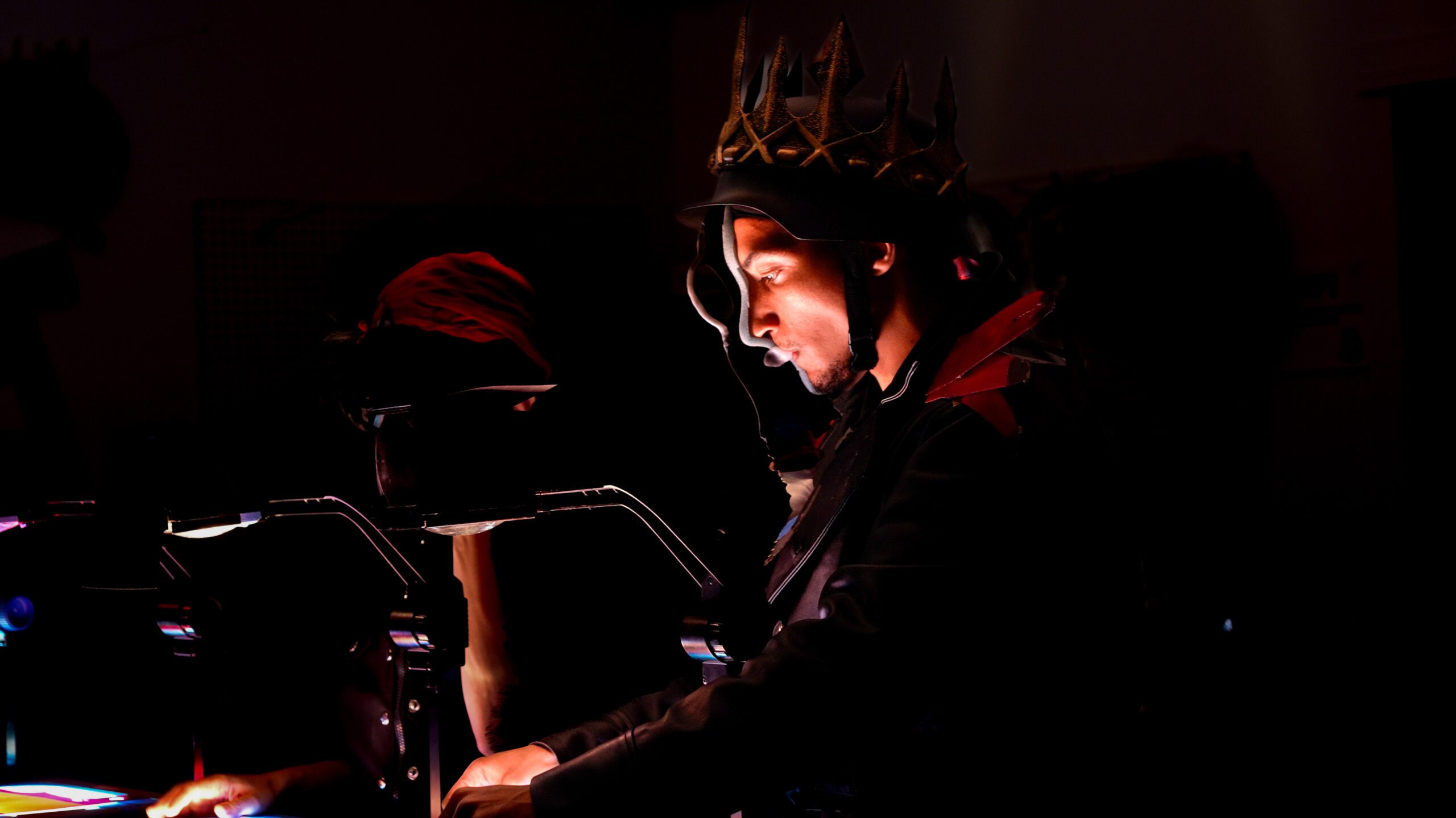 A person wearing a dark costume with a spiked crown stands at a lit table, focused on their hands. Another person, mostly in shadow, is nearby. The scene is dramatic with low lighting and vivid highlights.