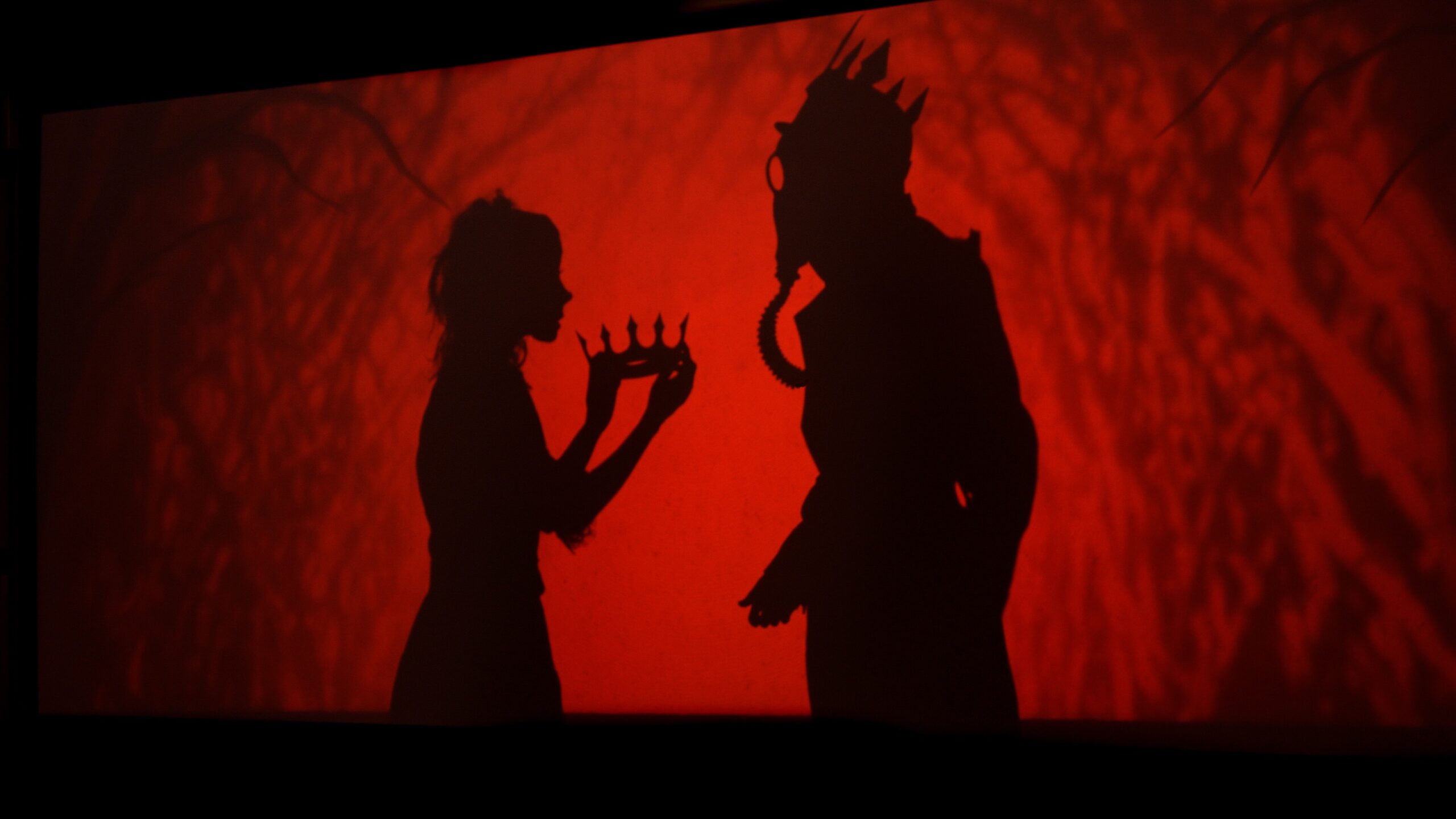 Two people’s silhouettes appear on a red background. One figure, possibly female, holds a crown, while the other wears a spiked crown and a gas mask. The scene is dramatic and theatrical.