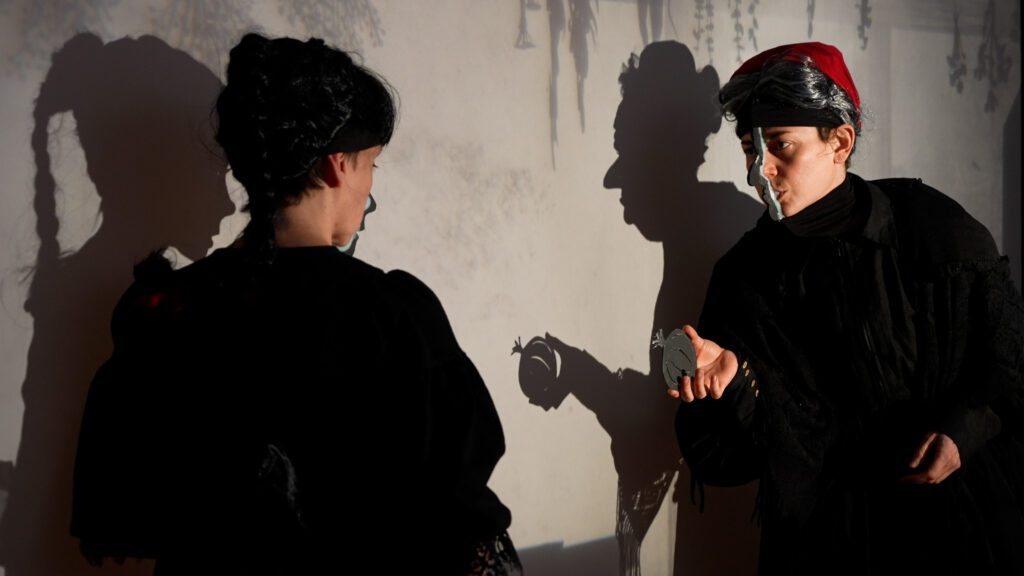 Two people in dark costumes face each other, casting dramatic shadows on a wall. One person extends a hand, holding an object, while both shadows add an intense atmosphere to the scene.