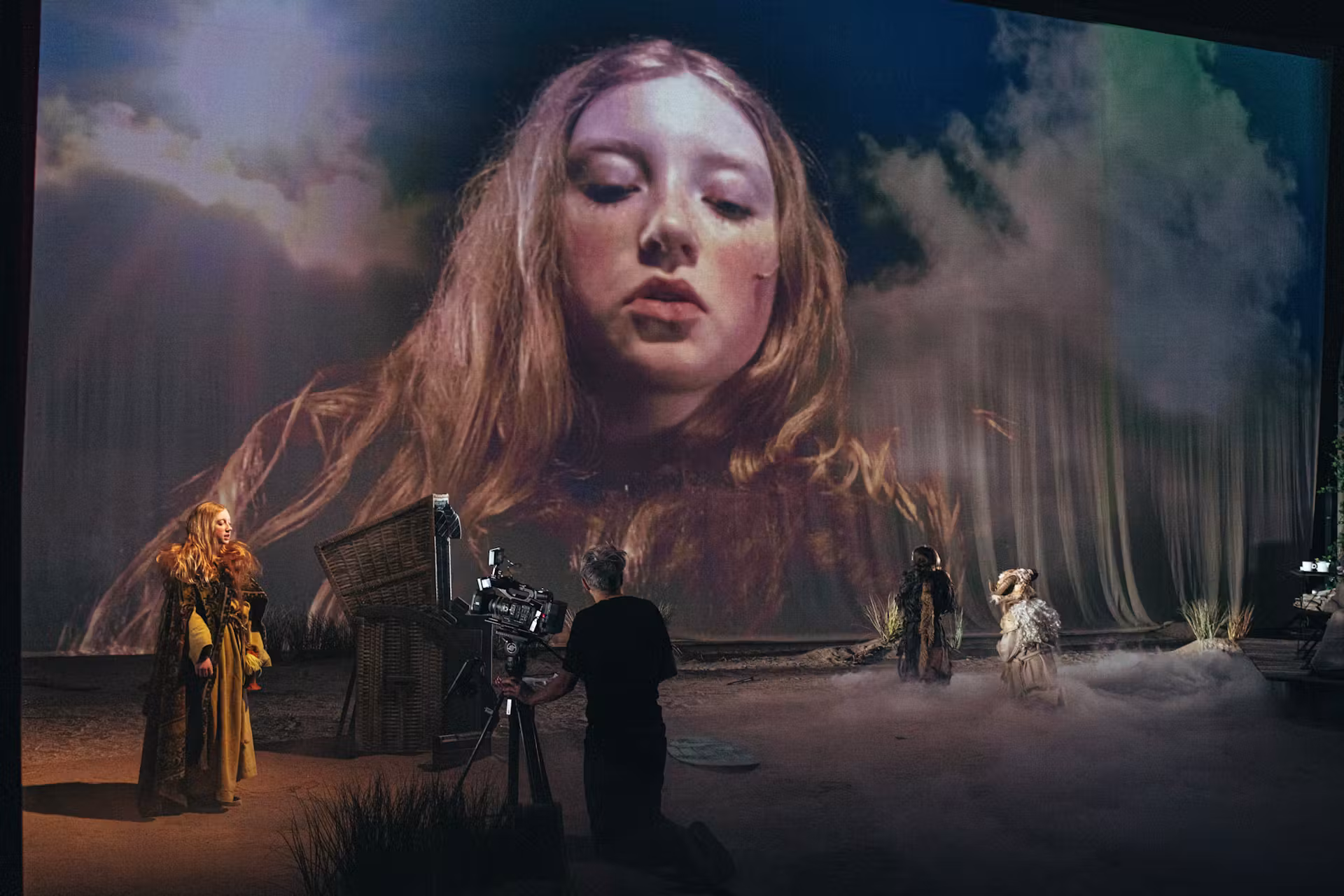 A film crew records actors in costume on a foggy set, while a giant projection of a woman’s face with long hair appears in the background, creating a surreal, dramatic scene.