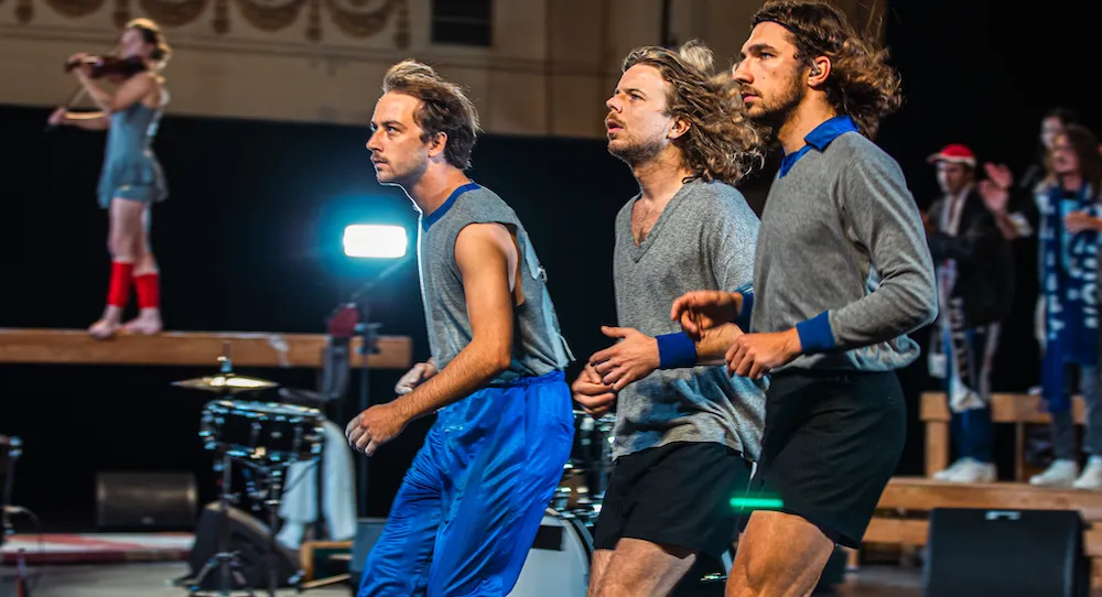 Three men in athletic clothing jog indoors, focused ahead. A woman in the background stands on a platform playing violin. Other people and musical equipment are visible in the scene.