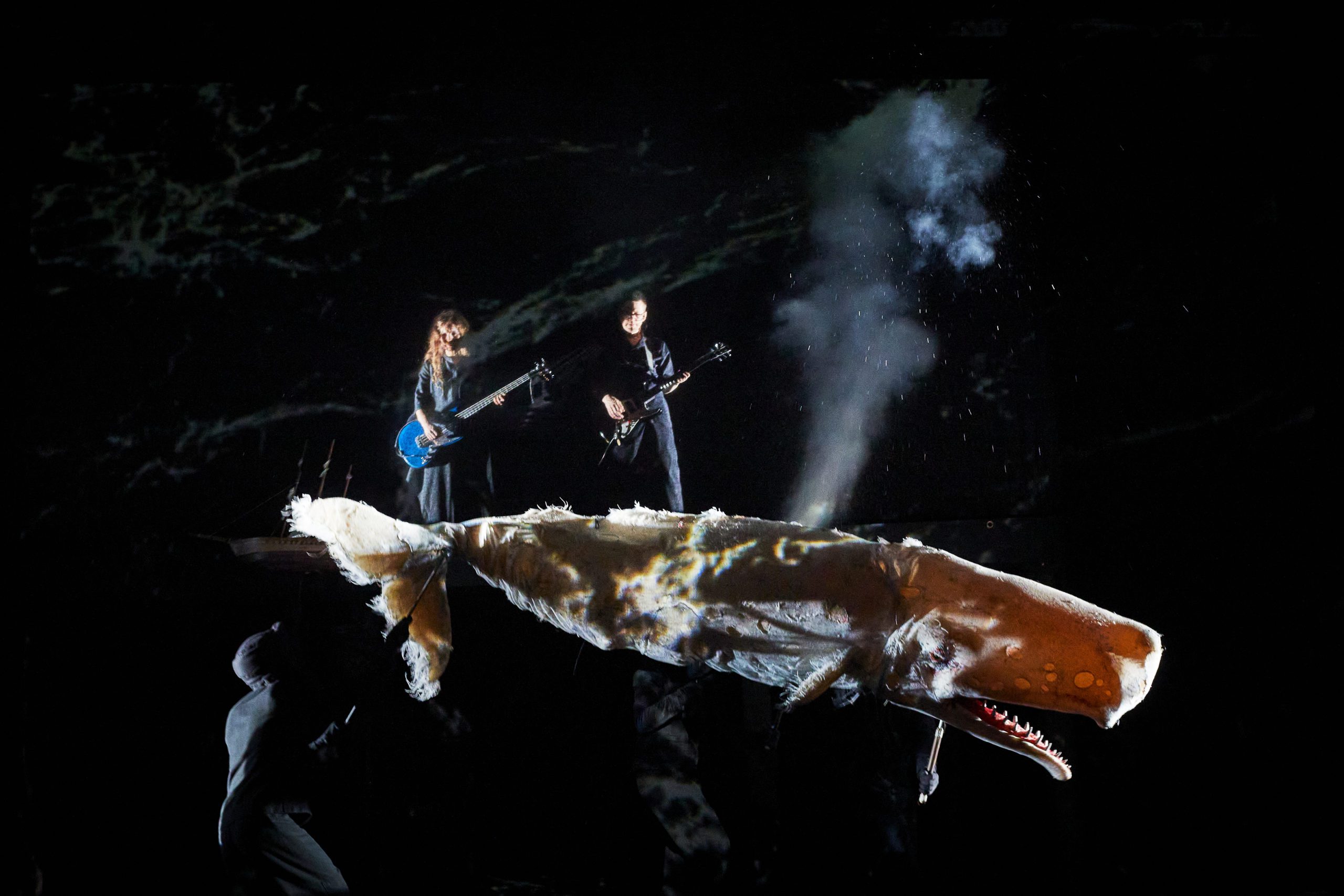 Two musicians with guitars perform on stage behind a large, whale-shaped puppet that appears to be spouting mist, with dark lighting and dramatic shadows.
