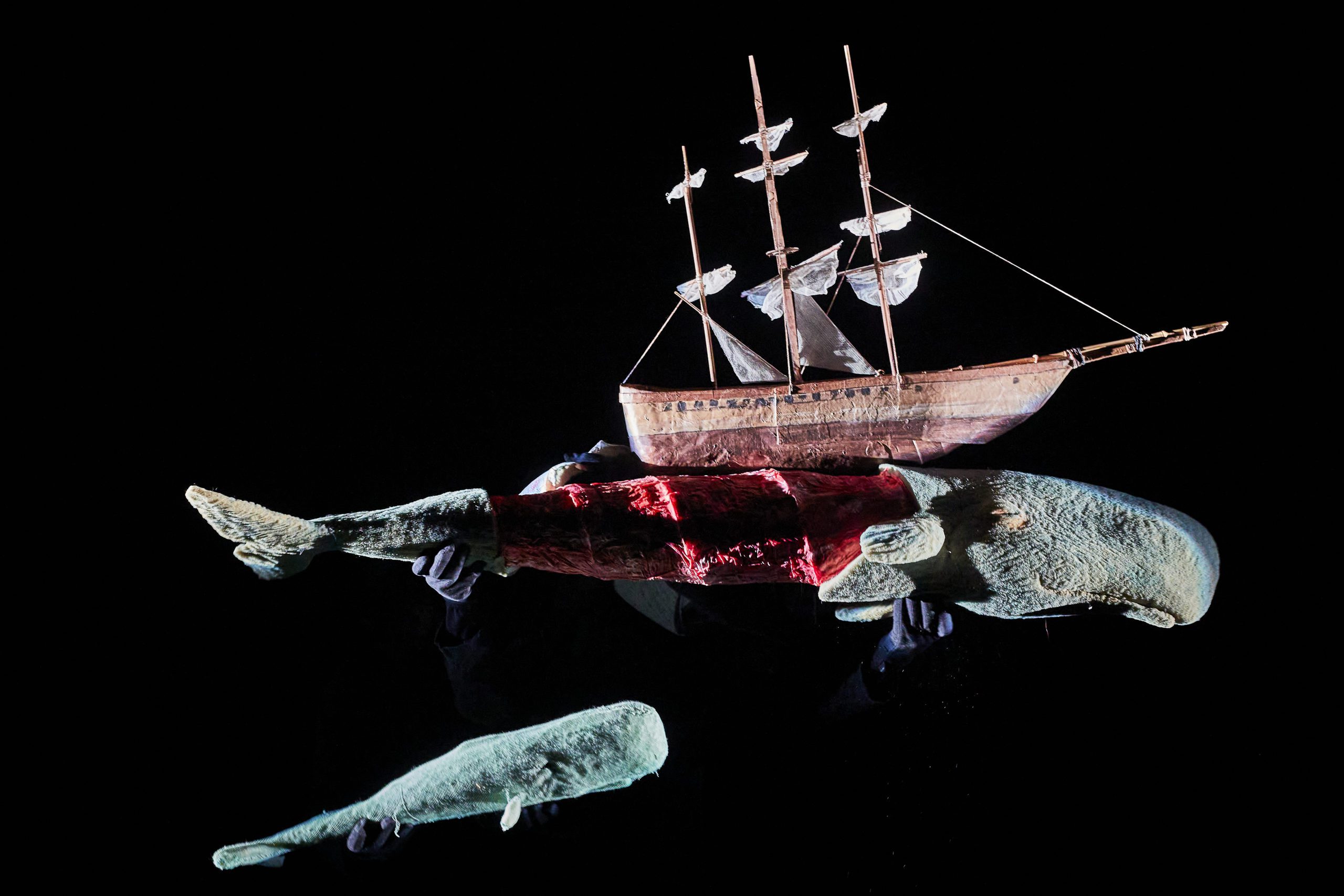Three puppets—a wooden ship, a sliced-open whale, and a smaller whale—are held against a black background by barely visible gloved hands.
