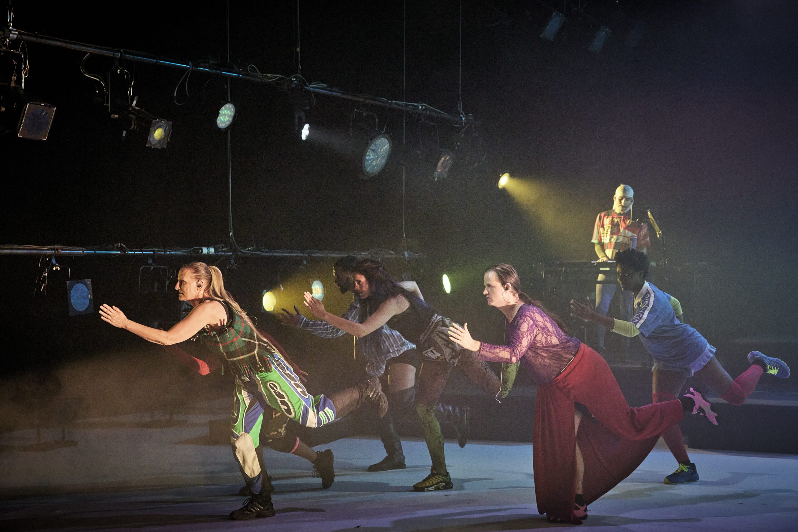 Four performers in colorful, eclectic costumes pose as if running forward on stage, with dramatic lighting above. In the background, a DJ stands at a console, partially illuminated.