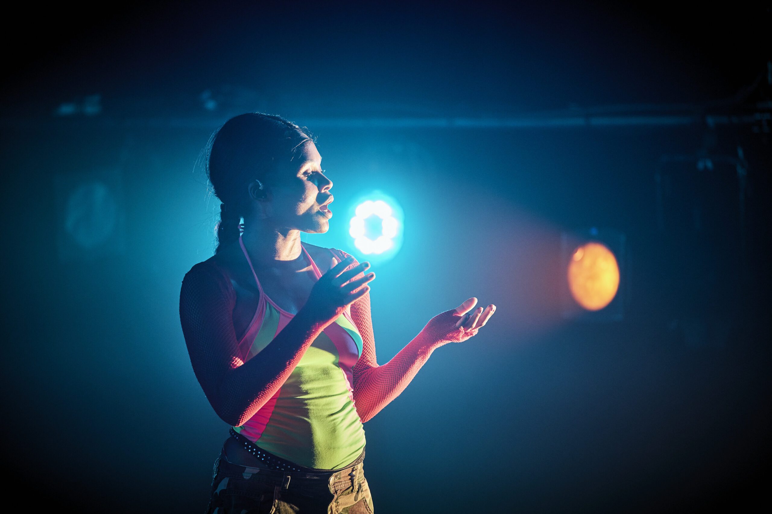 A person in colorful clothing stands under dramatic stage lighting, illuminated by blue and orange spotlights, with one hand raised and a thoughtful expression.
