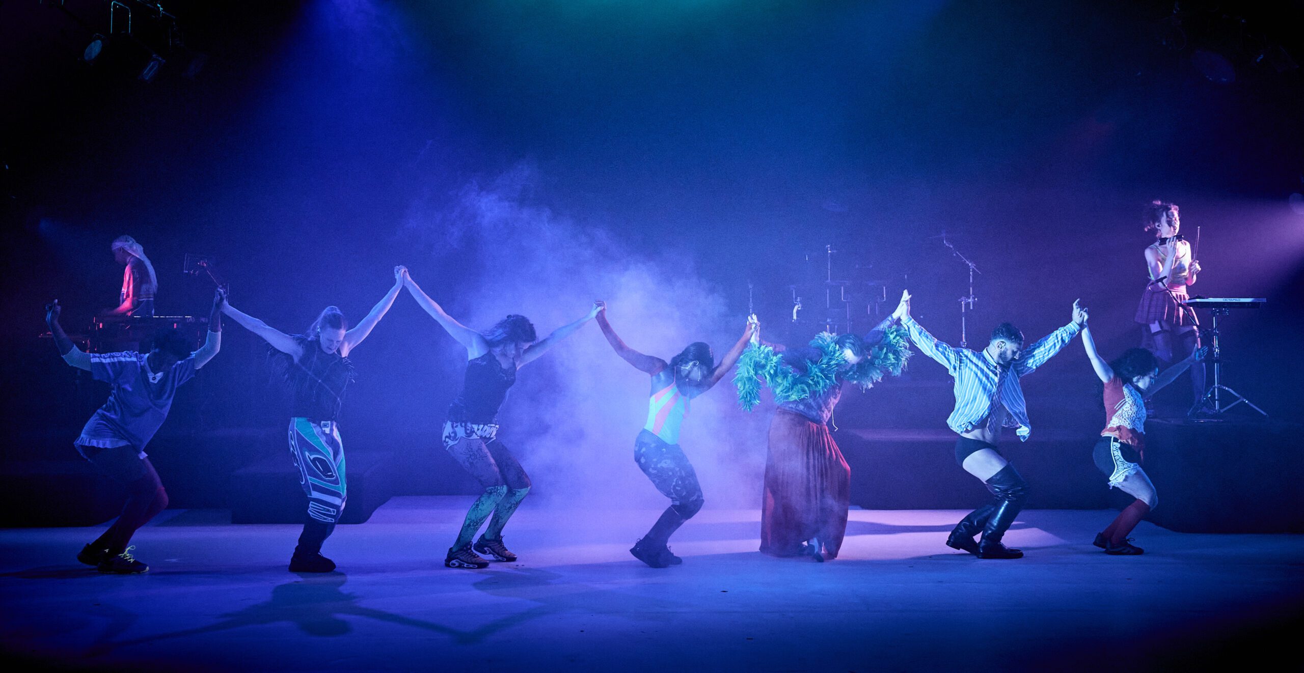 Seven performers hold hands and dance in a line under colorful stage lights, with smoke in the background. Two musicians play keyboards at opposite sides of the stage.