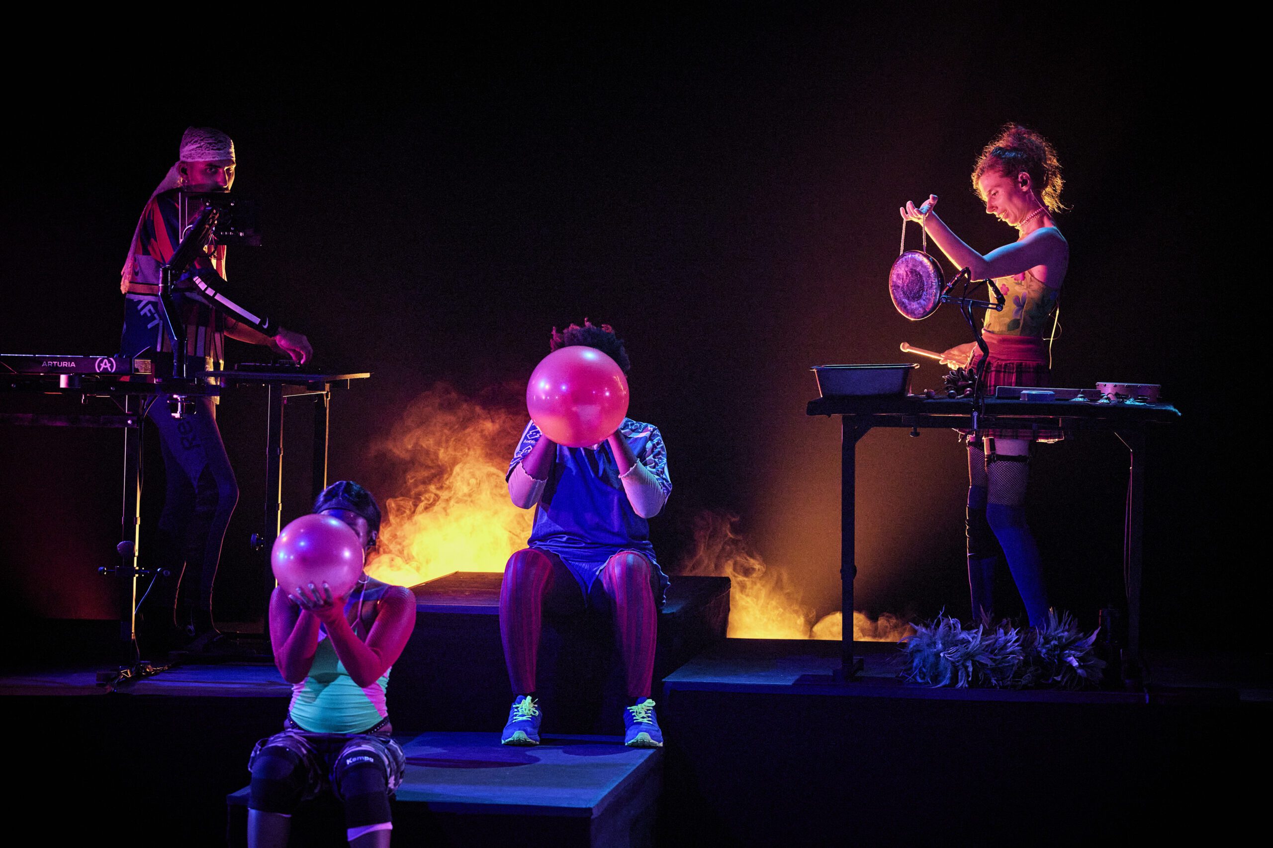 Four performers are on stage with colorful lighting and smoke. Two people are inflating pink balloons, while another plays a synth and the fourth strikes a metallic gong.