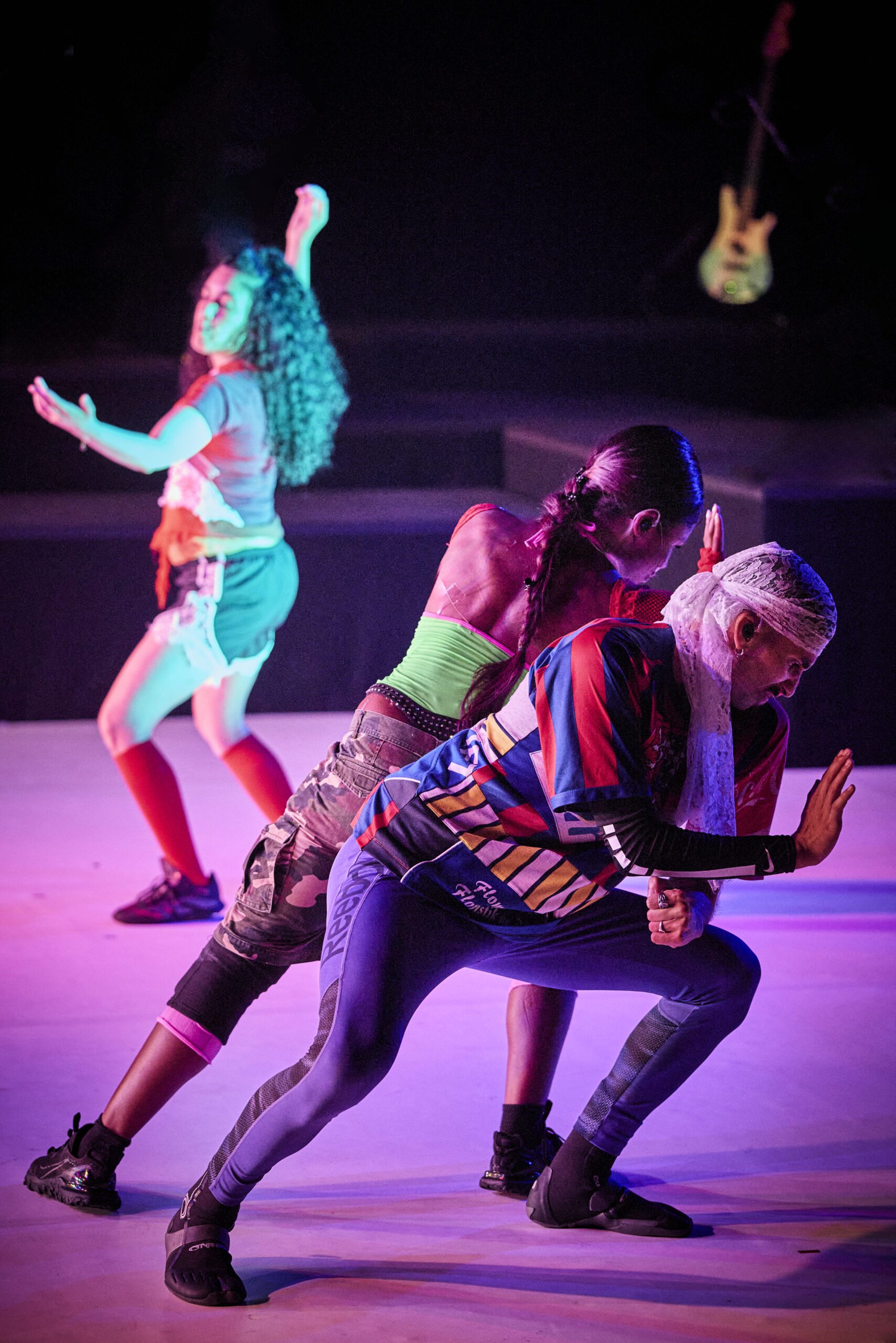 Three dancers pose on stage under colorful lights, wearing athletic clothes. Two face opposite directions, back-to-back, while the third dances in the background. Their movements appear expressive and synchronized.