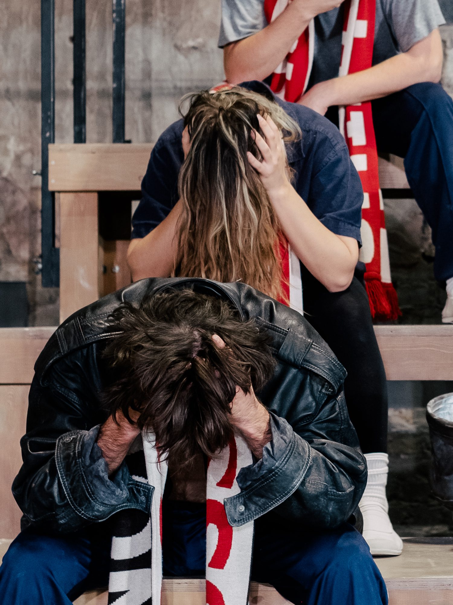 Two people sit on bleachers with their heads in their hands, looking distressed. Both wear red and white scarves, and the person in front is wearing a black jacket.