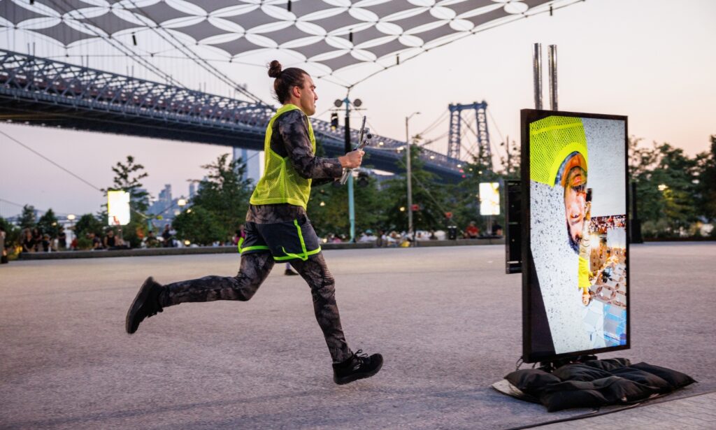 A person in athletic clothing runs outdoors near a bridge, passing a screen displaying their image with digital effects. Trees and people are visible in the background during sunset.