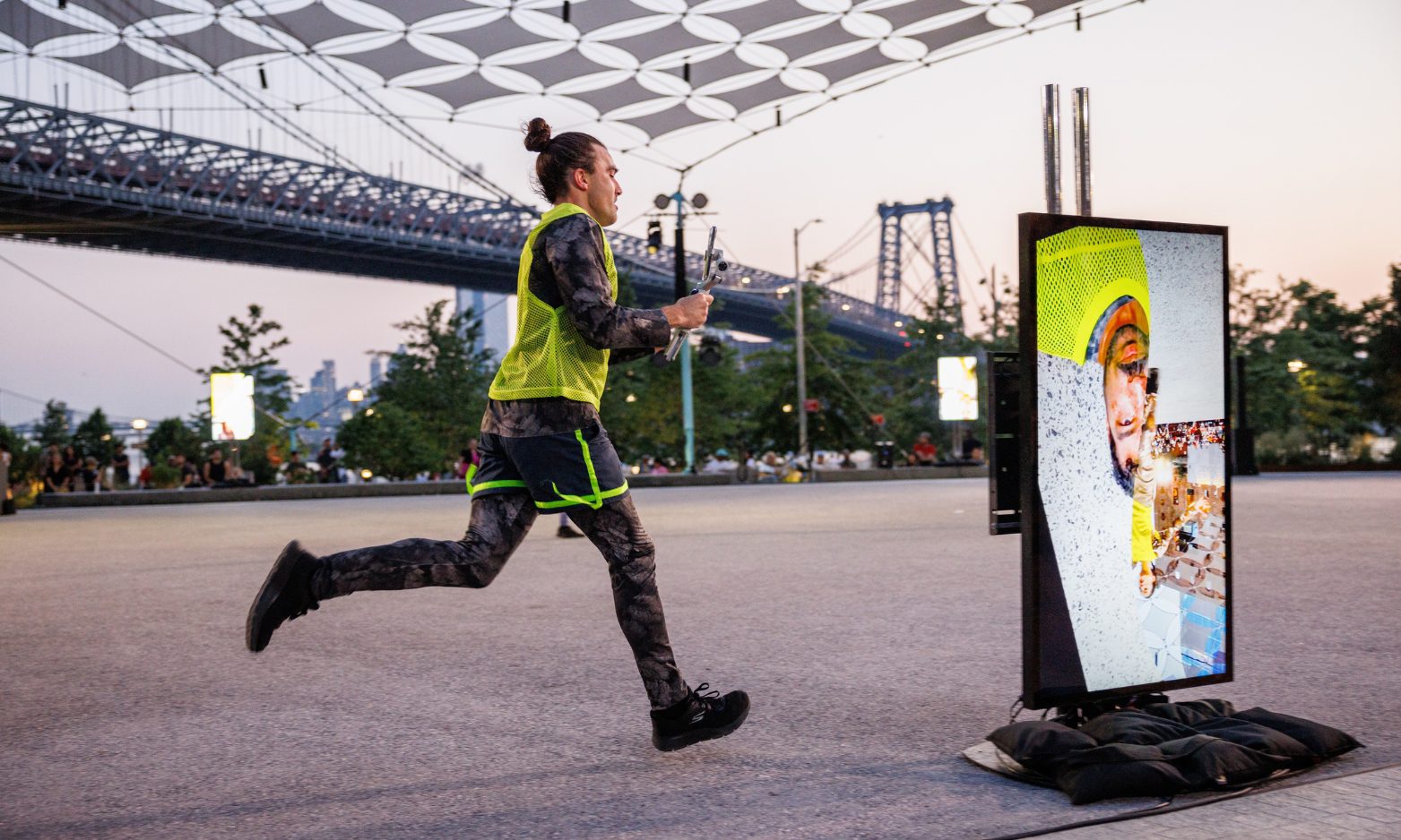 A person in athletic clothing runs outdoors near a bridge, passing a screen displaying their image with digital effects. Trees and people are visible in the background during sunset.
