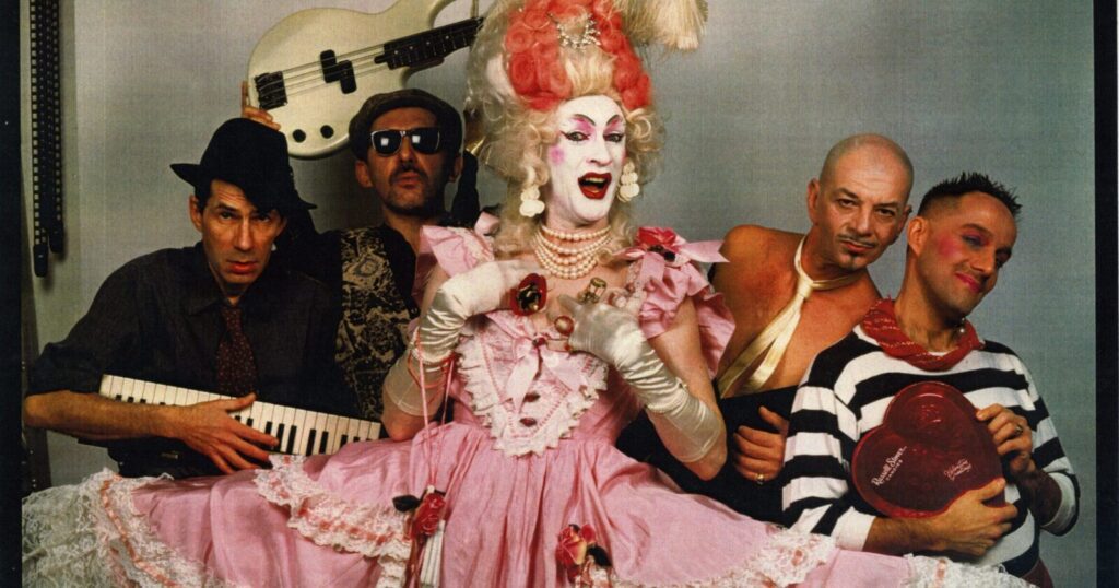 Five people pose together; the central figure wears an elaborate pink dress, white gloves, and dramatic makeup. Others wear eclectic costumes and hold musical instruments, including a keyboard, guitar, and bass guitar.