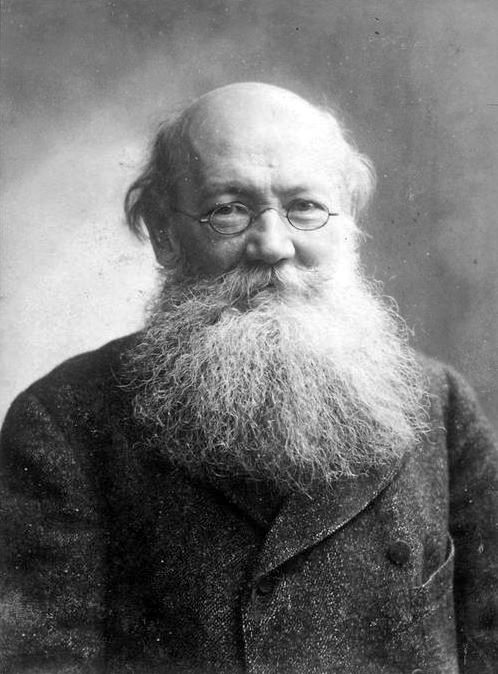 An older man with a large white beard, glasses, and a receding hairline wearing a dark coat, looking directly at the camera in a black-and-white portrait.