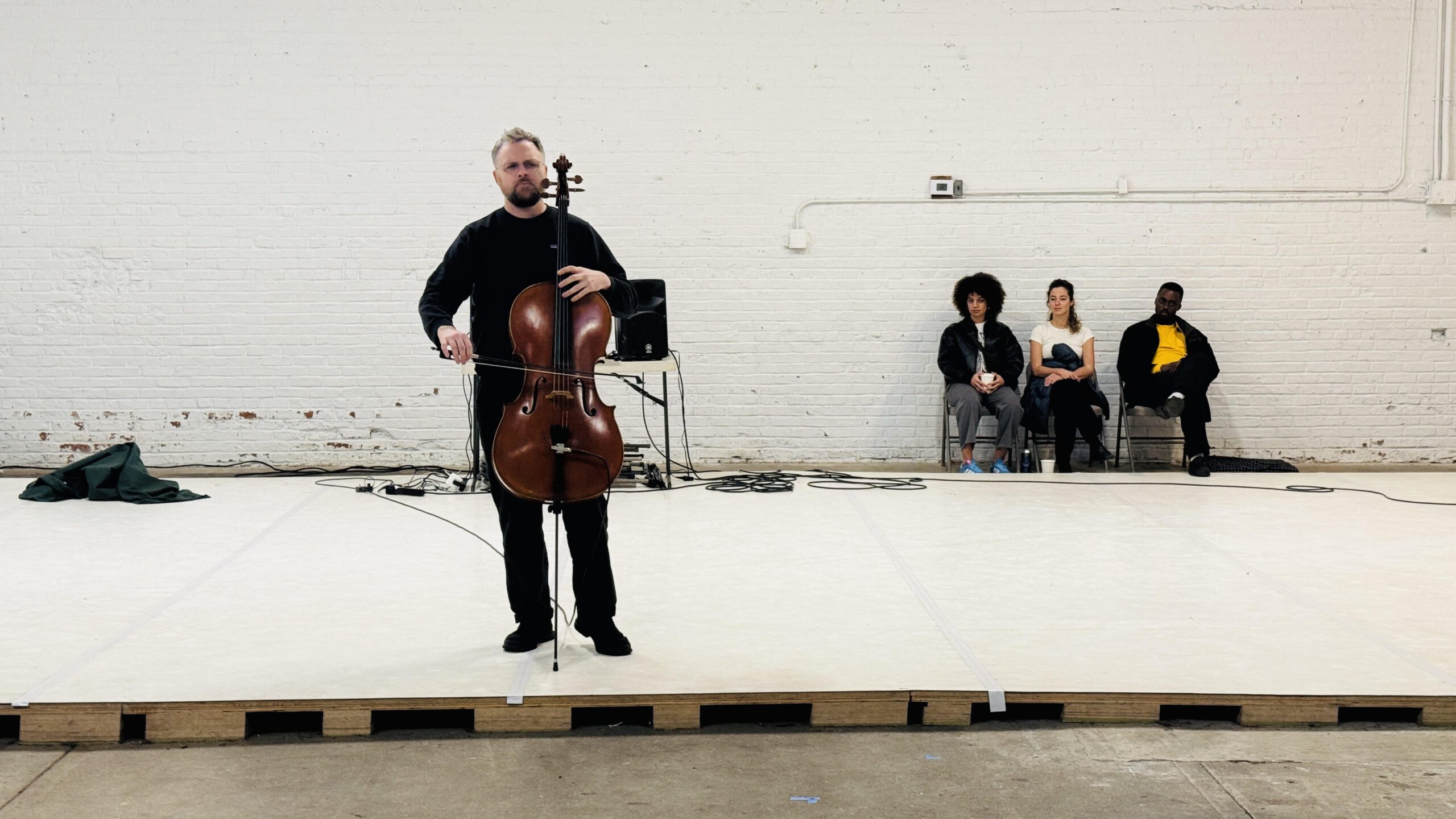 A cellist in black performs on a platform in a large, industrial-style room. Three people sit in the background against a white brick wall, watching.