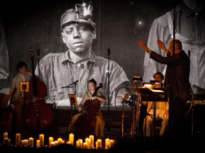 Musicians perform on stage with a conductor, illuminated by candles, in front of a large projected black-and-white image of a miner in work clothes and a headlamp.