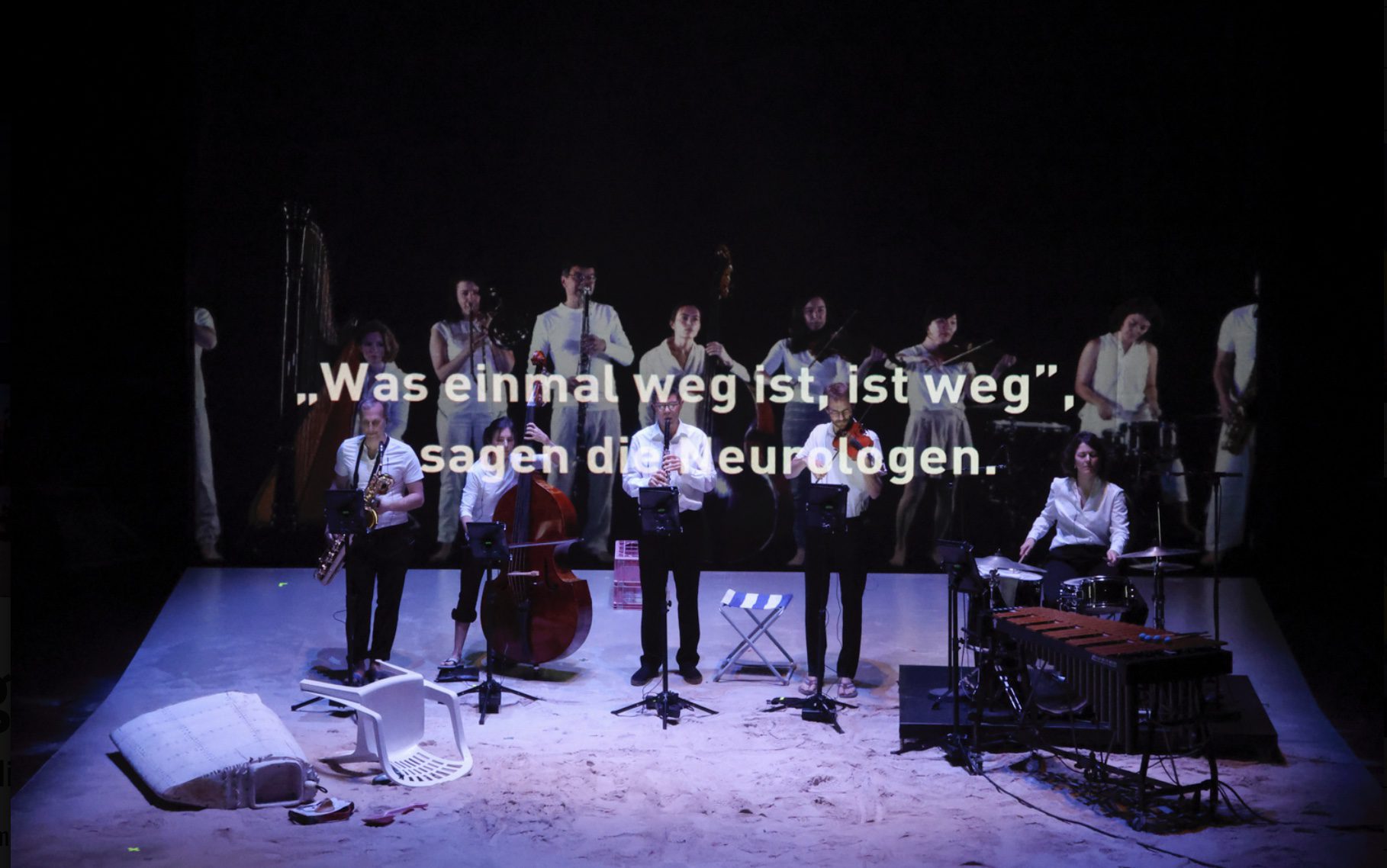 A group of musicians perform onstage with instruments; German text on the image reads: “Was einmal weg ist, ist weg,” sagen die Neurologen. The stage is dimly lit with scattered objects in the foreground.